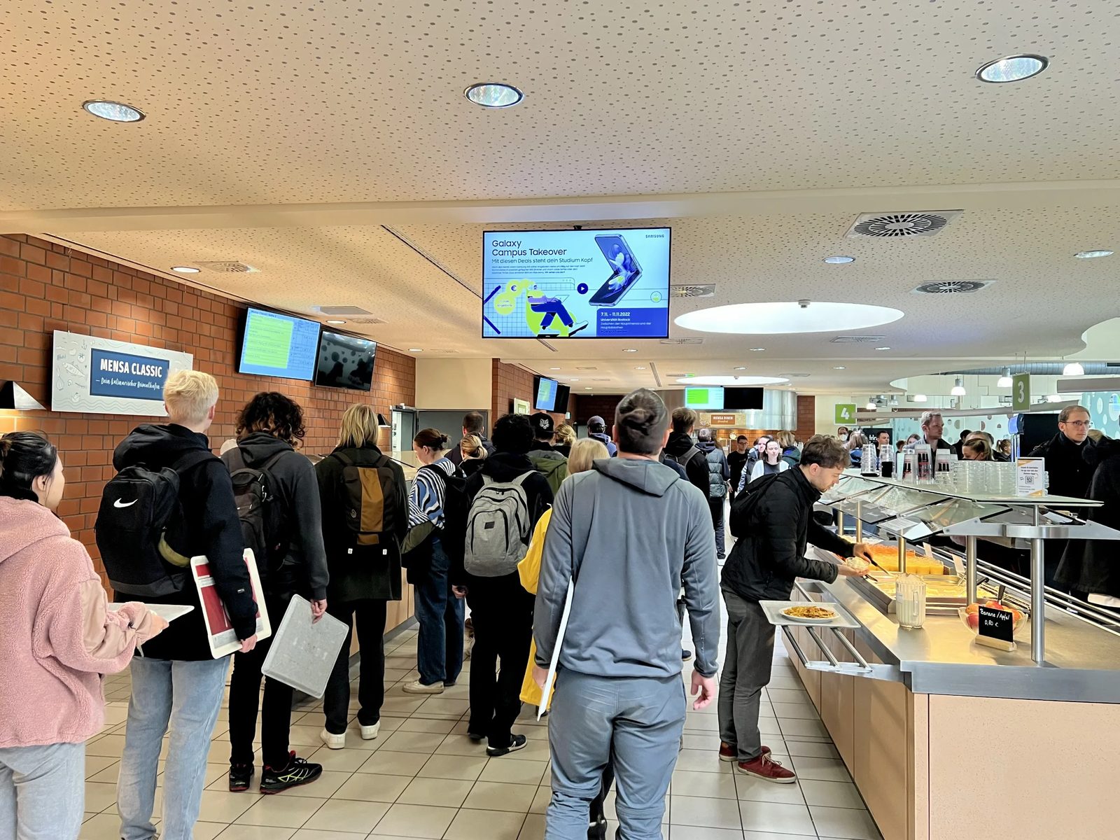 Students queuing in a university cafeteria under ceiling screens showing the menu and a campus offer