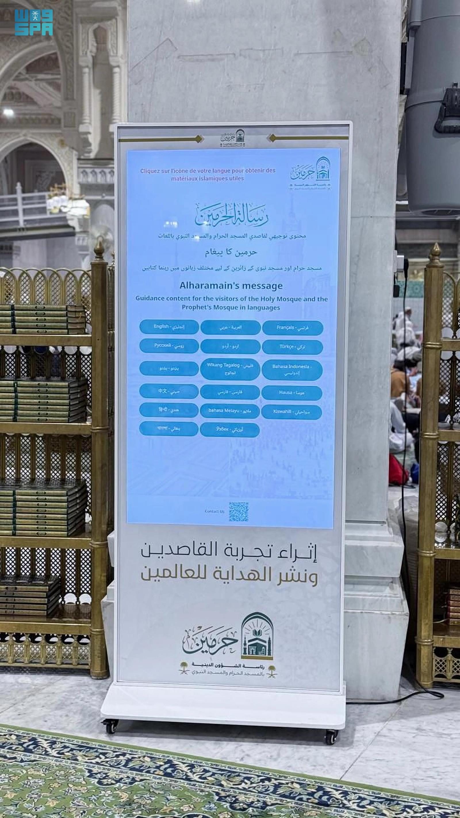 A tall standing screen in a mosque showing guidance for visitors in many languages, beside shelves of Qurans