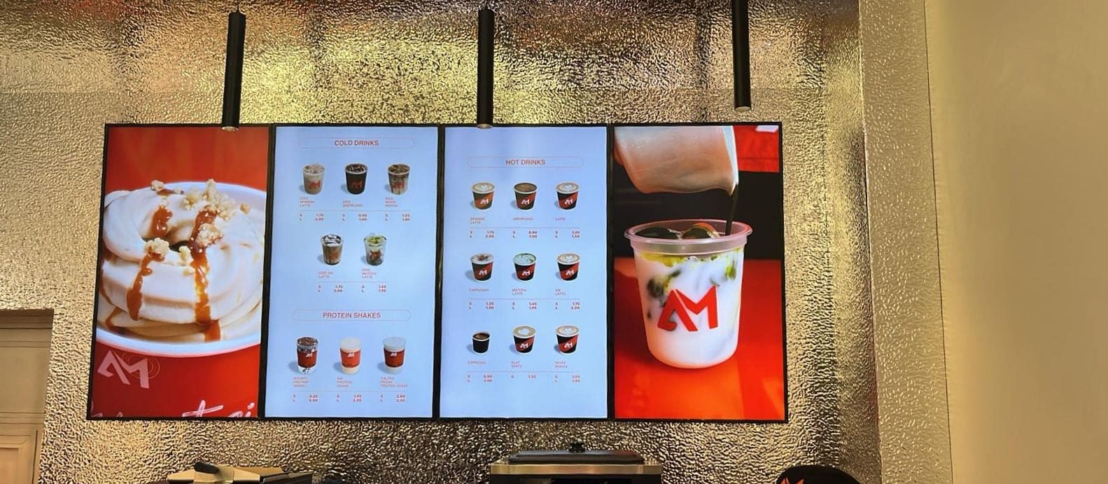 Four screens side by side on a cafe wall, two showing drinks menus and two showing photos of a dessert and a drink