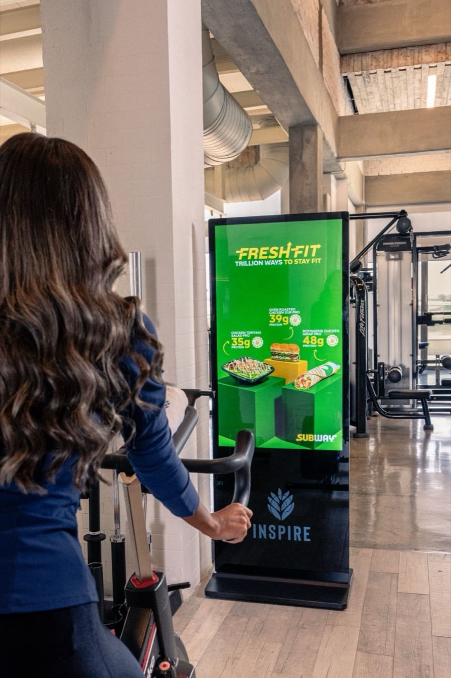 Subway's Fresh Fit ad on a standing screen beside a woman on an exercise bike, in a mixed gym in Kuwait