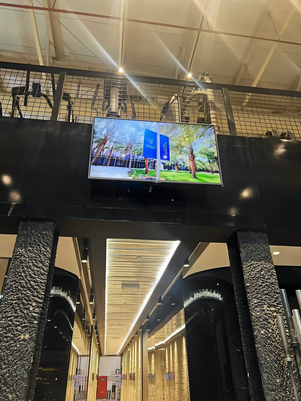 AIU's campus ad on a wall screen above the floor of a mixed gym, under the mezzanine's netting